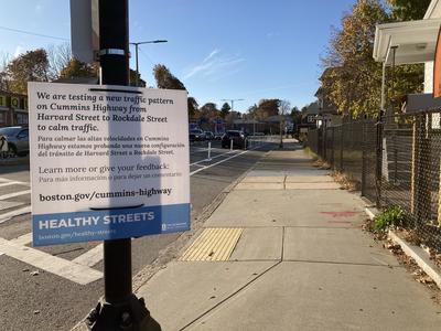 A sign is posted reading: We are testing a new traffic pattern on Cummins Highway from Harvard Street to Rockdale Street to calm traffic. In the background is a parking-protected bike lane.