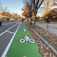 A green bike lane extends forward from view, with a car traveling to the left in a travel lane. Trees line the street to the right, and a person is walking on the sidewalk.