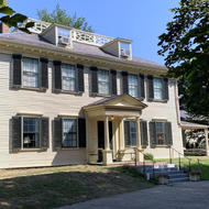 View of an 18th-century Georgian mansion with light yellow siding and dark green shutters. An ell extends off to the right.