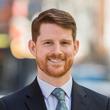 A photo of a man with red hair and a beard wearing a black suit jacket and a light blue tie.